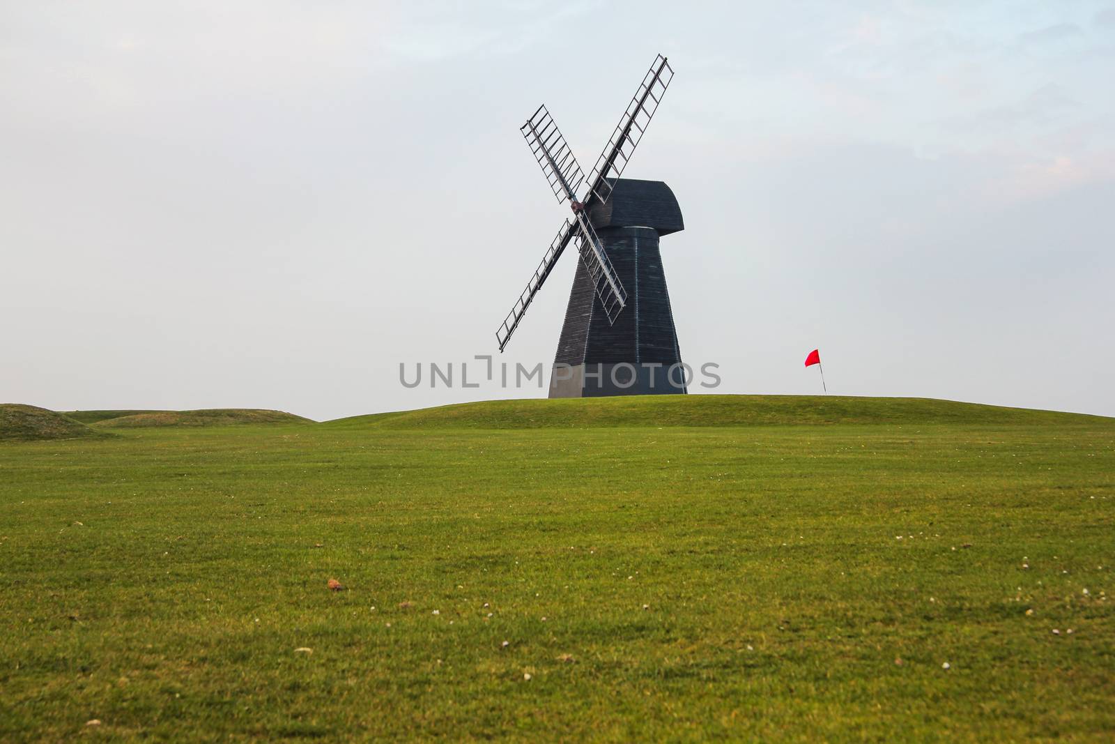 Windmill and red flag with green lawn and overcast sky in Rottin by Ivanko