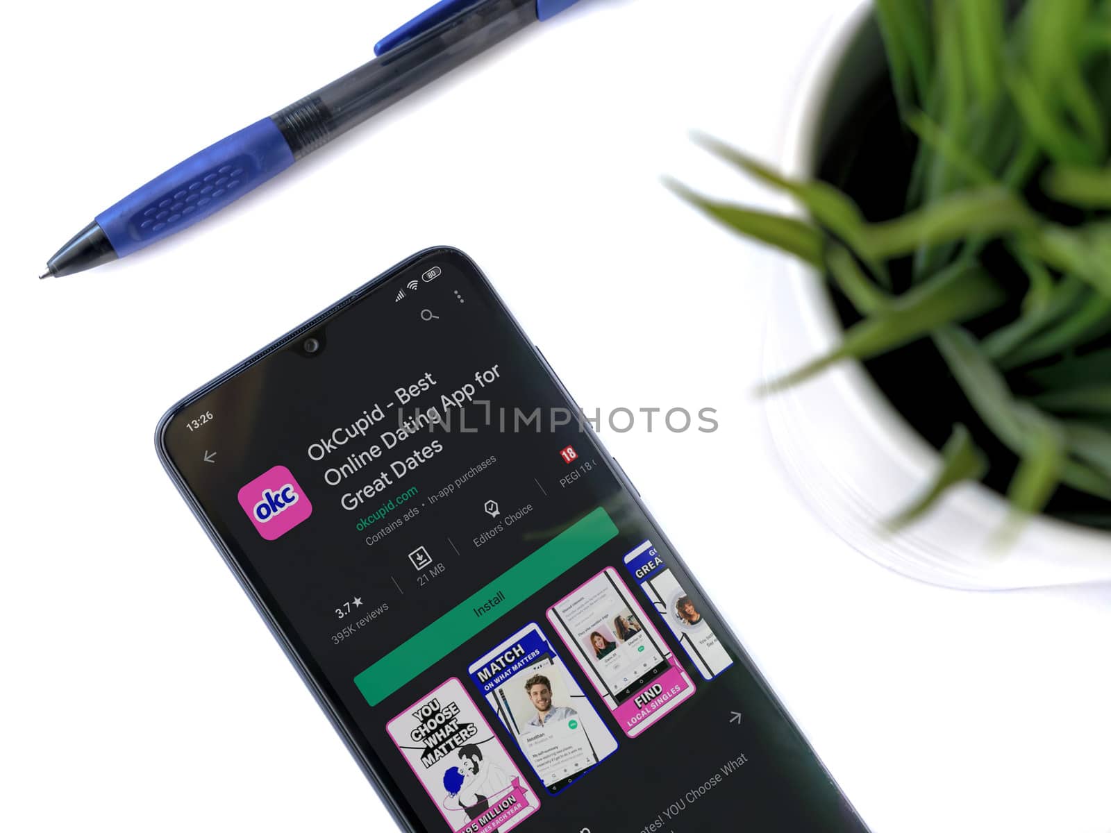 Lod, Israel - July 8, 2020: Modern minimalist office workspace with black mobile smartphone with OkCupid app play store page on a white background. Close up top view flat lay.