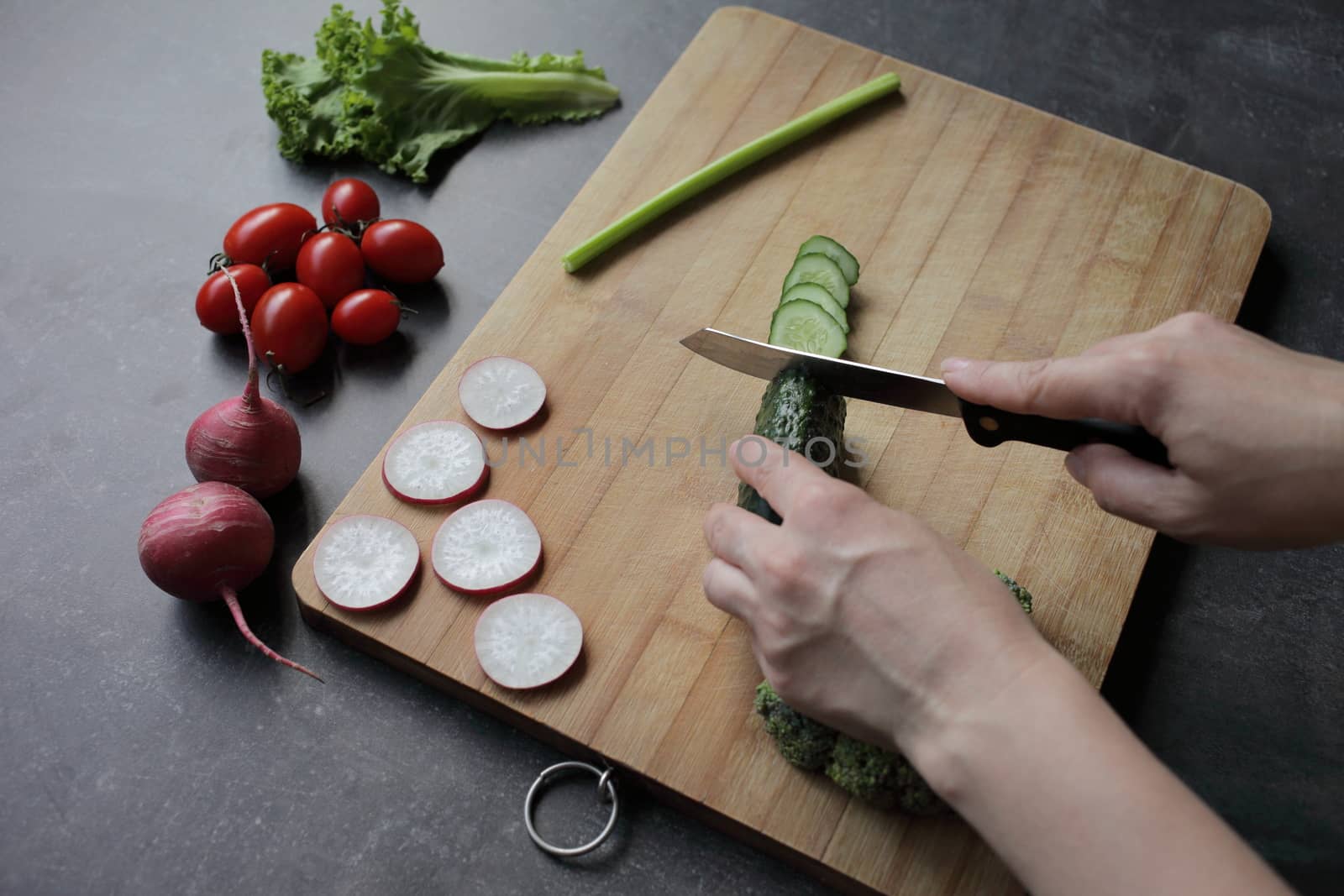 Hands cut cucumber on a cutting board on a gray table. Fresh vegetables, tomatoes, lettuce, broccoli, radishes lie nearby