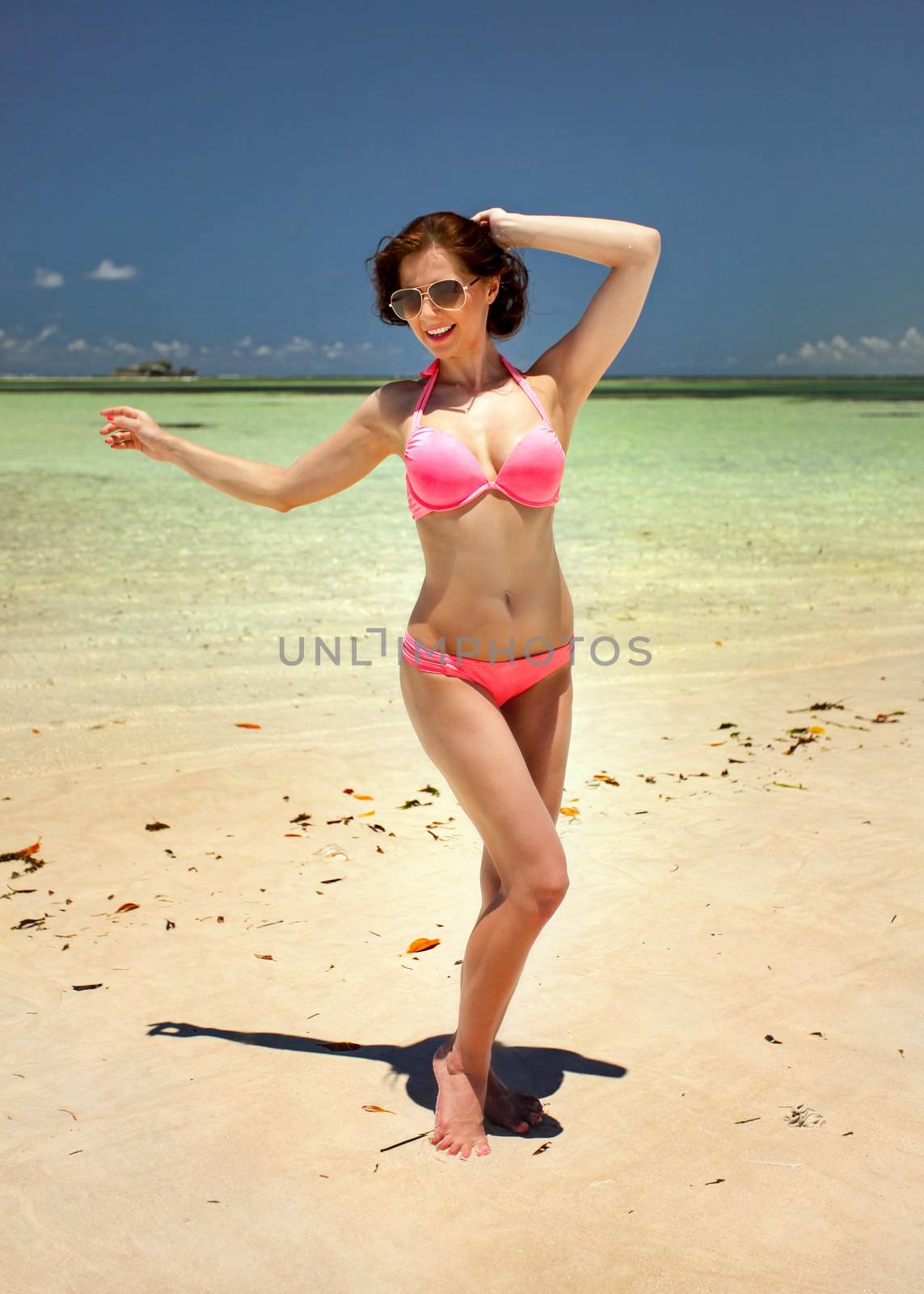 Sexy young woman with sunglasses holding her dark hair posing as if dancing on fine sand beach with calm clear sea in background. Watamu, Kenya