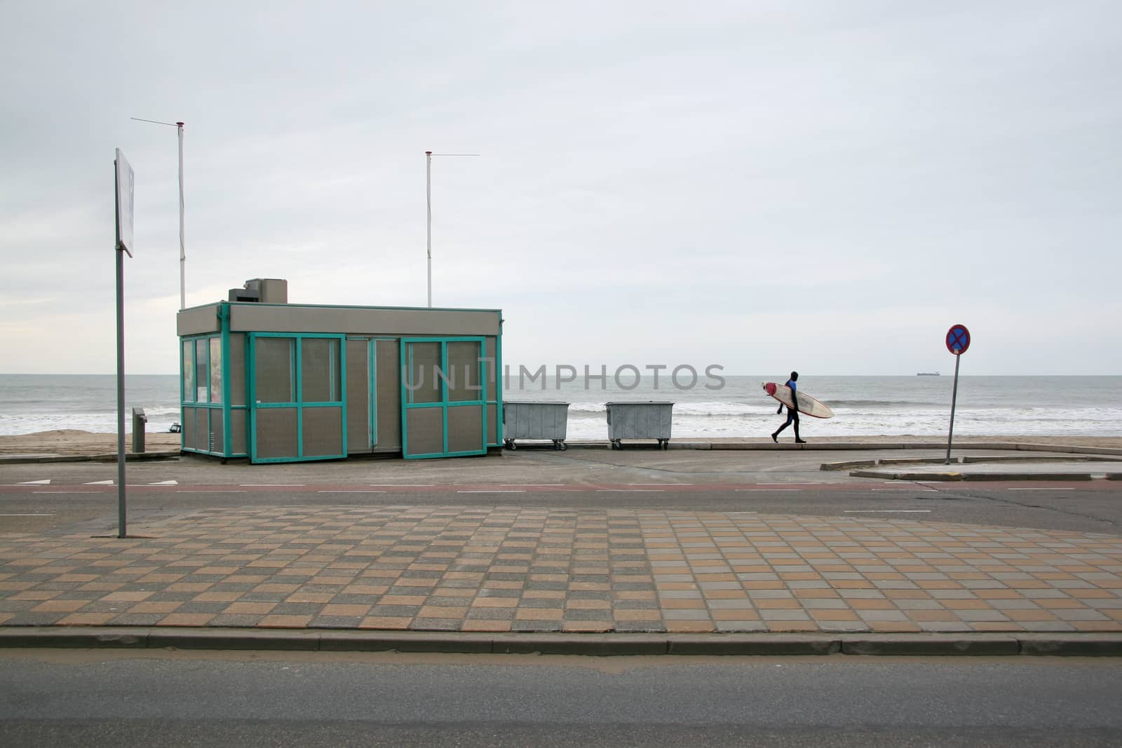 Lone surfer in neoprene swimsuit walking on pavement to empty beach on a cold winter overcast day. The Hague, Netherlands