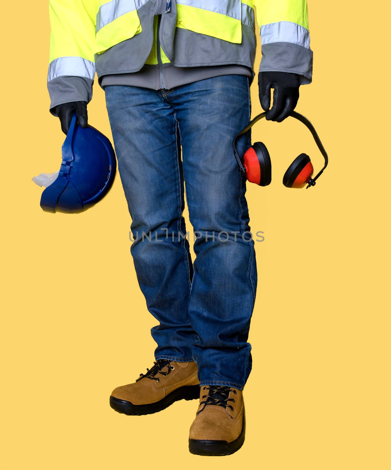 Workman wearing jeans, gloves, boots and holding protective equipment, helmet and ear defender on yellow background
