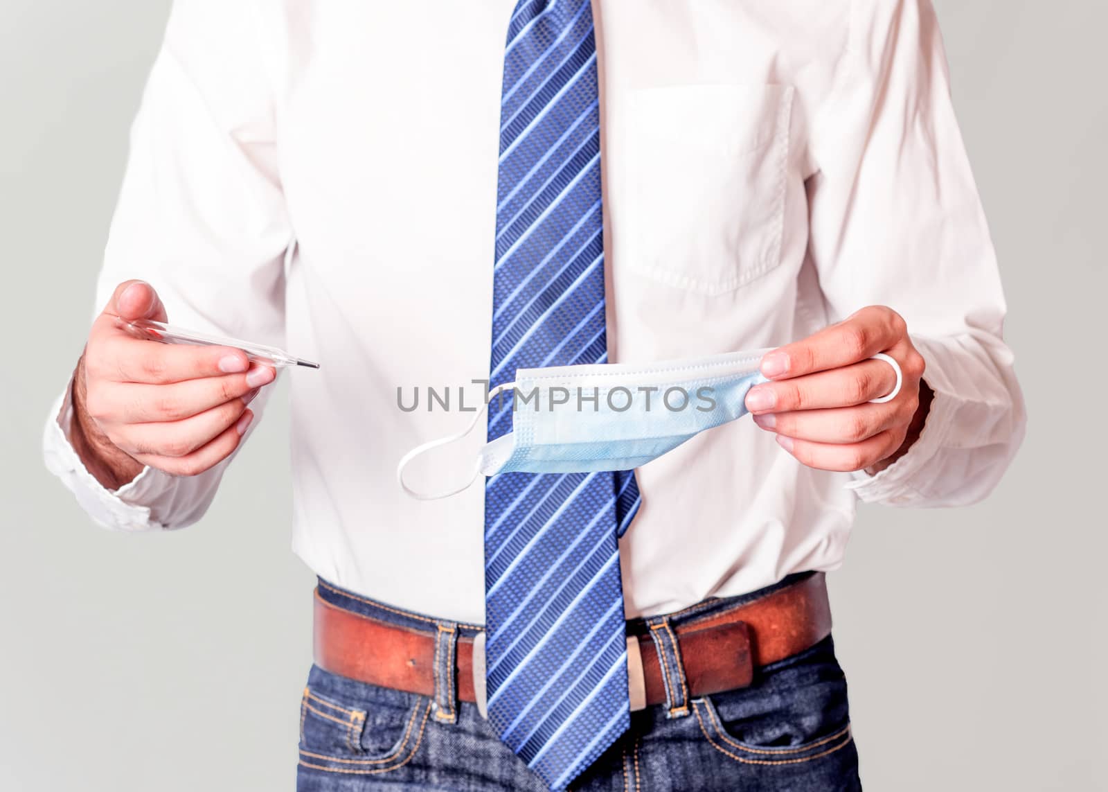 man in white shirt and jeans holding face mask and thermometer on grey background