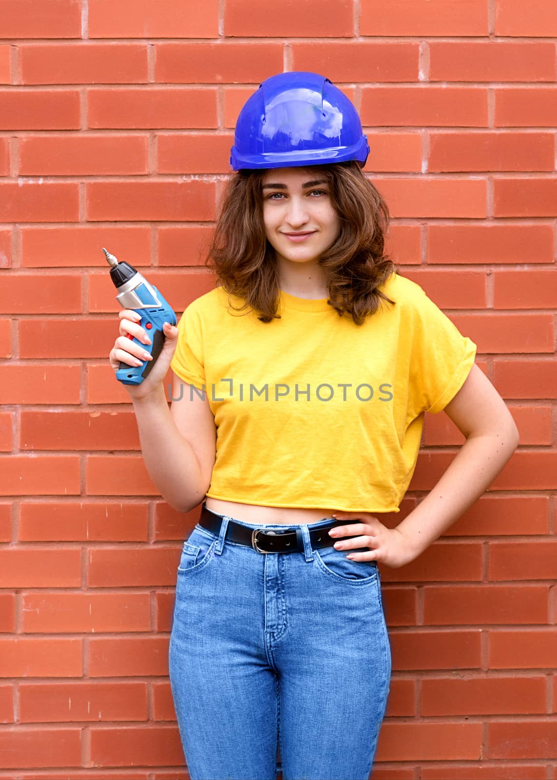 a female construction worker in yellow shirt and blue helmet with a screwdriver against brick wall