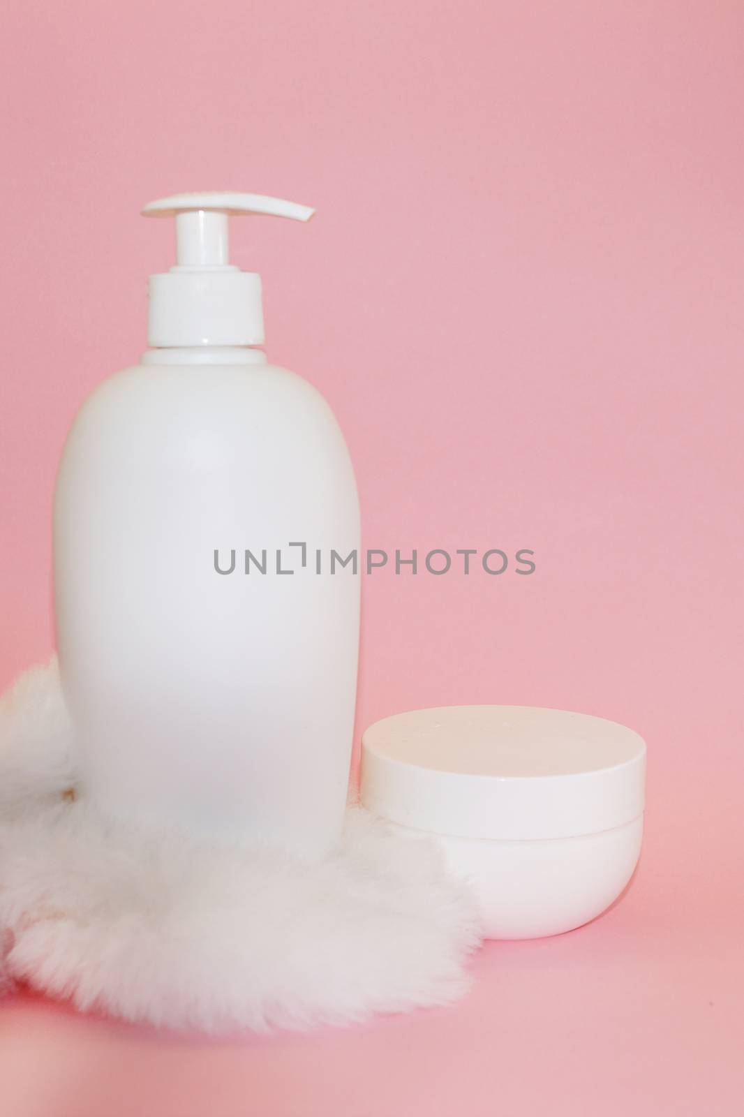 white cosmetic bottle, cream jar and white fur on pink background close-up