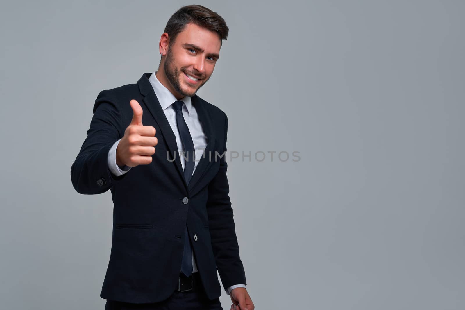 Portrait young smiling businessman. Caucasian guy business suit tie studio gray background. Modern business person Showing thumbs up sign. Portrait of charming successful happy entrepreneur
