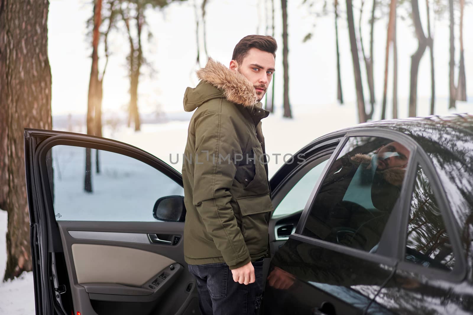 Man stands winter forest road car. Holiday trip. Cold season. Portrait young handsome unshaved driver. Modern hipster guy traveling on car