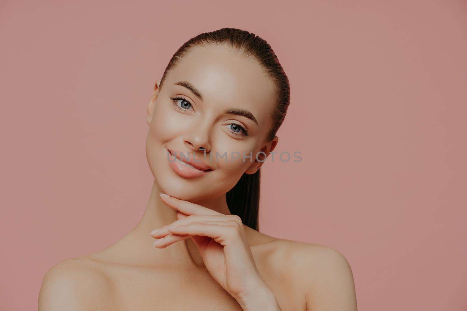 Charming beautiful woman with pony tail, touches perfect soft skin after cosmetology procedures, using beauty cream, stands with bare shouldes, poses against pink background. Natural as she is by vkstock