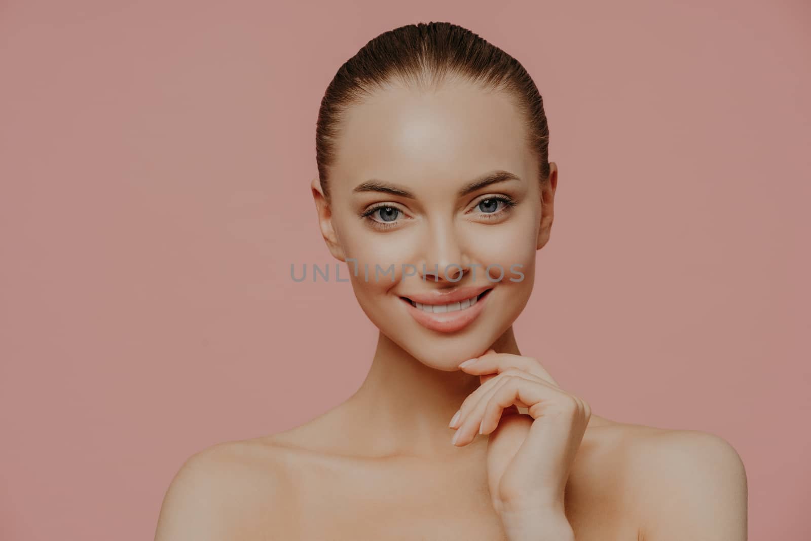 Studio shot of pretty charming woman touches chin, has healthy glowing skin, cares about complexion, stands naked indoor, has cosmetology procedure, isolated on pink background. Beauty concept