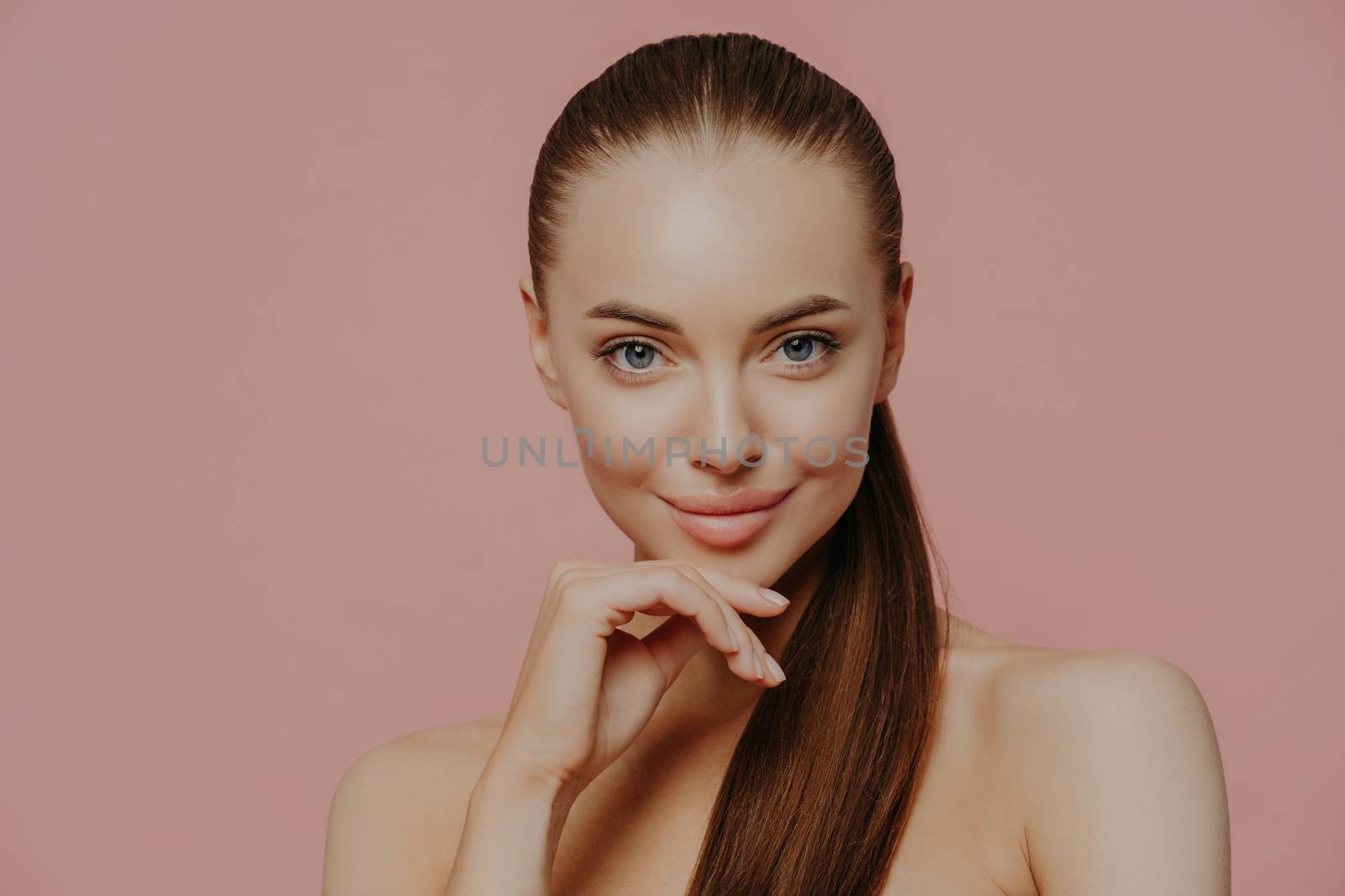 Skin care and anti aging procedures. Confident brunette young woman keeps hand under chin, has combed hair, clean fresh skin, enjoys beauty treatments, poses with naked body over pink background by vkstock