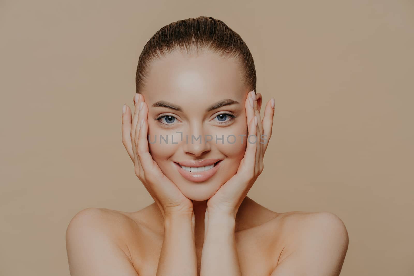 Skin care and beauty concept. Headshot of beautiful cheerful woman touches face gently, has perfect smile, healthy skin after cleaning or applying facial mask, isolated over beige background