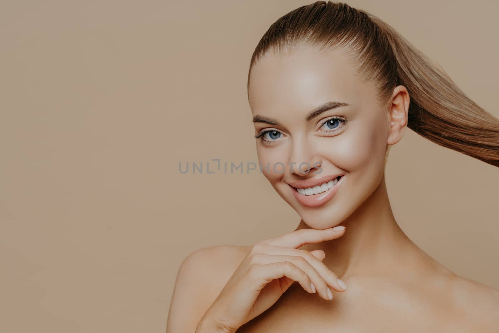 Tranquil undressed woman with healthy perfect skin, dark hair combed in pony tail, enjoys facial treatments, has well cared complexion, stands against beige studio wall, empty space. Skin care concept by vkstock
