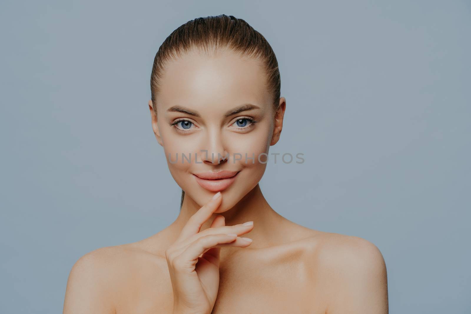 Tender sensual woman with pure healthy skin, has natural magnetic look, feels beautiful and attractive, stands shirtless against blue background, enjoys herself after spa procedures. Beauty care by vkstock