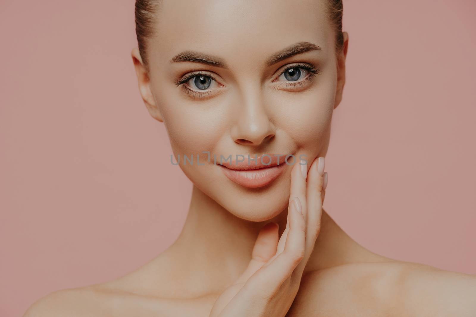 Cropped shot of attractive calm woman touches face gently, has direct gaze at camera, cares about skin, has well groomed skin, enjoys facial skin care, stands naked against pink studio background