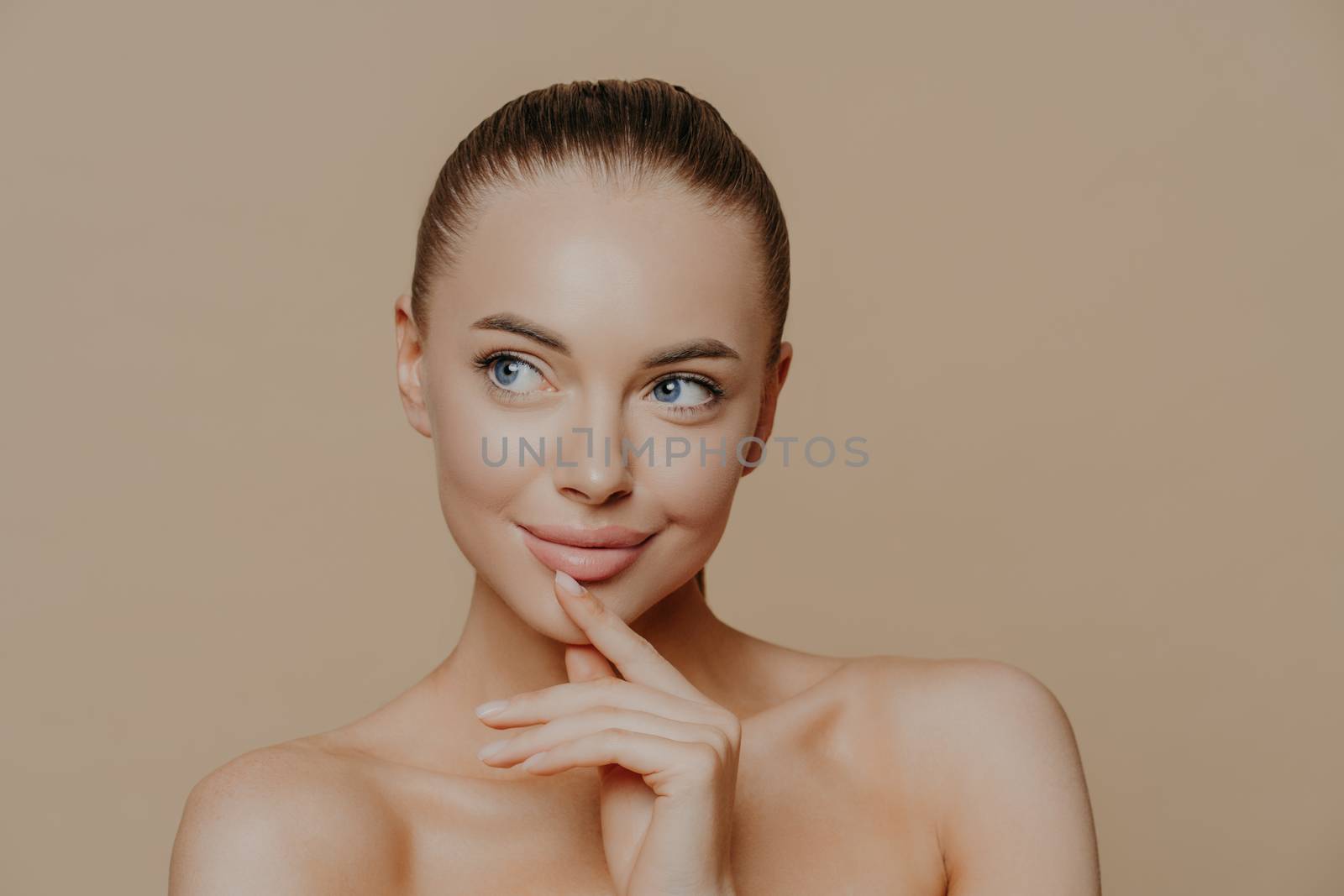 Pensive attractive young European woman with white healthy skin, looks aside, has naked body, undergoes beauty treatments, poses against beige background, has combed dark hair, bare shoulders