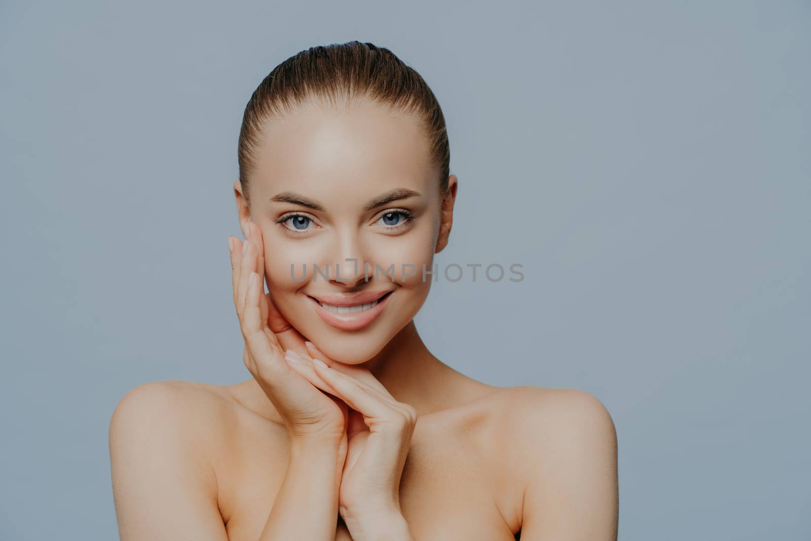 Studio shot of pretty smiling woman touches face and enjoys flawless of skin, stands bare shoulders, has big forehead, poses against blue background, applies skin care cosmetics. Tenderness, makeup
