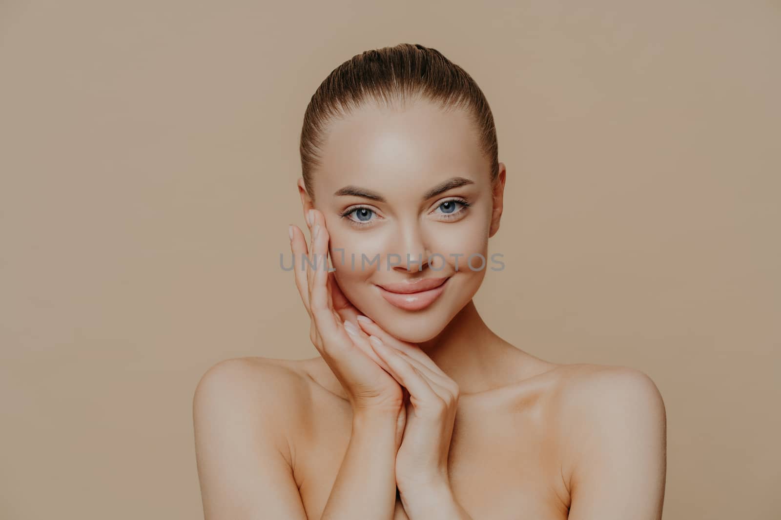 Natural beauty portrait. Photo of young woman enjoys perfect fresh skin after visiting cosmetologist, smiles gently, poses naked against beige background, touches face, wears minimal makeup.