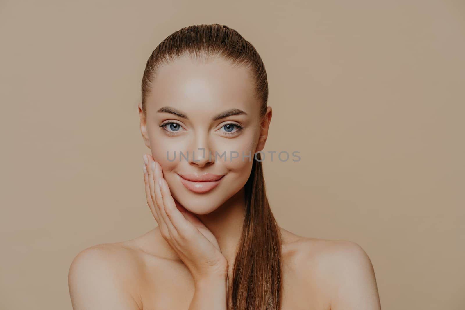 Photo of lovely young woman has smooth healthy skin after beauty procedures and receiving facial massage, stands bare shoulders, has long dark straight hair, isolated over beige wall cares of her body by vkstock