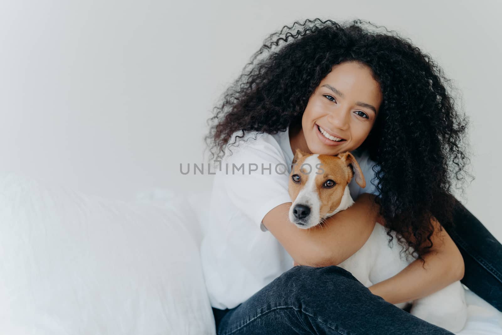 Photo of attractive young woman with Afro hircut, embraces with love dog, takes care of pet, smiles gently, wears casual clothing, isolated over white background, sit on bed, copy space for your promo by vkstock
