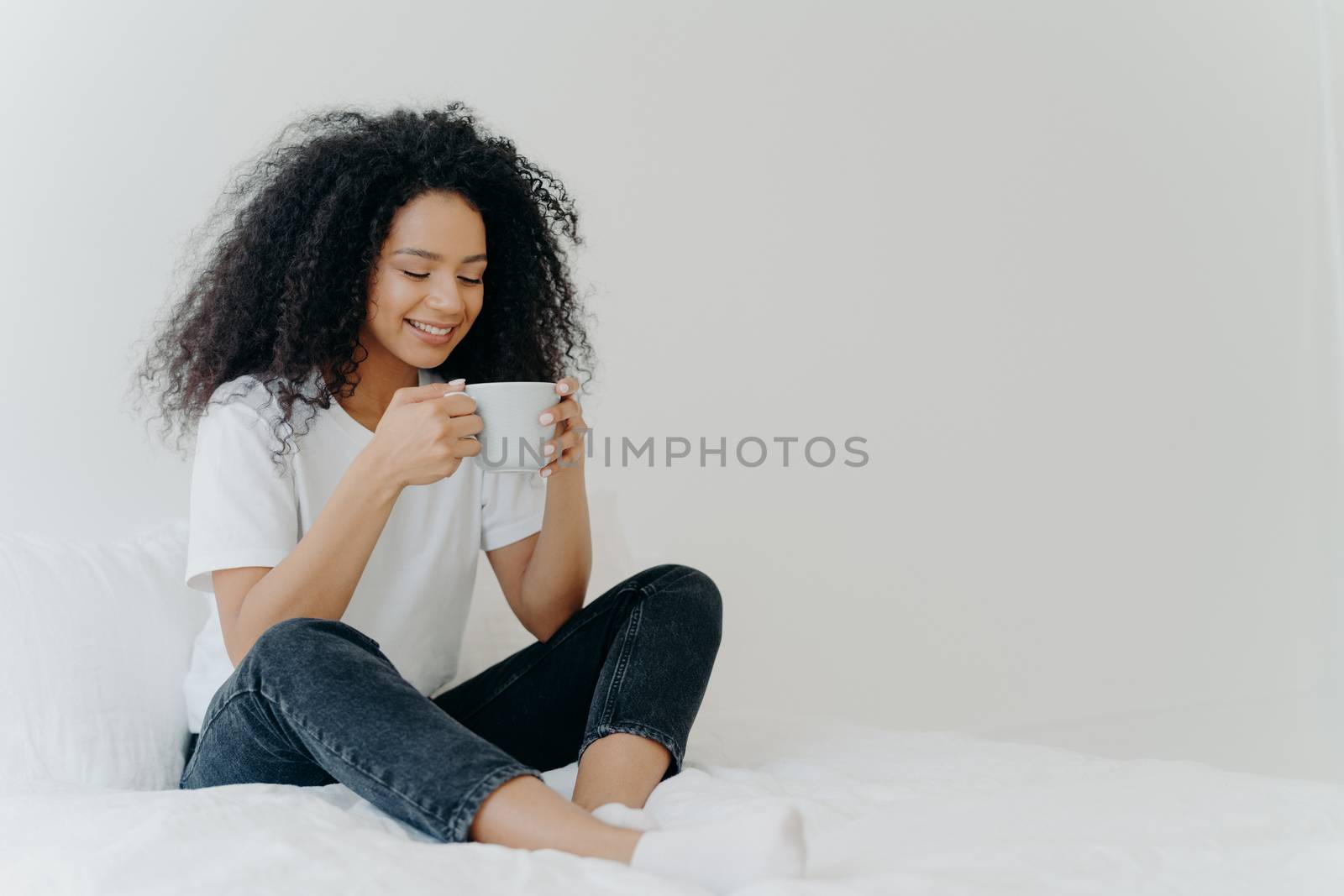 African American woman sips hot drink, poses in bed, enjoys good morning, dressed casually, spends weekend at home, blank space aside for your advertising. People, coziness and rest concept. by vkstock