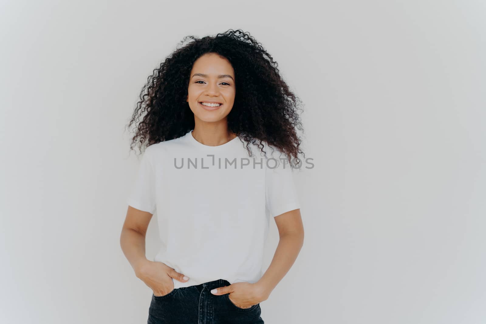 Isolated shot of young African American woman wears white t shirt, expresses good emotions, stands alone indoor, poses for photo, has casual talk with friend, enjoys free time. People and happiness by vkstock