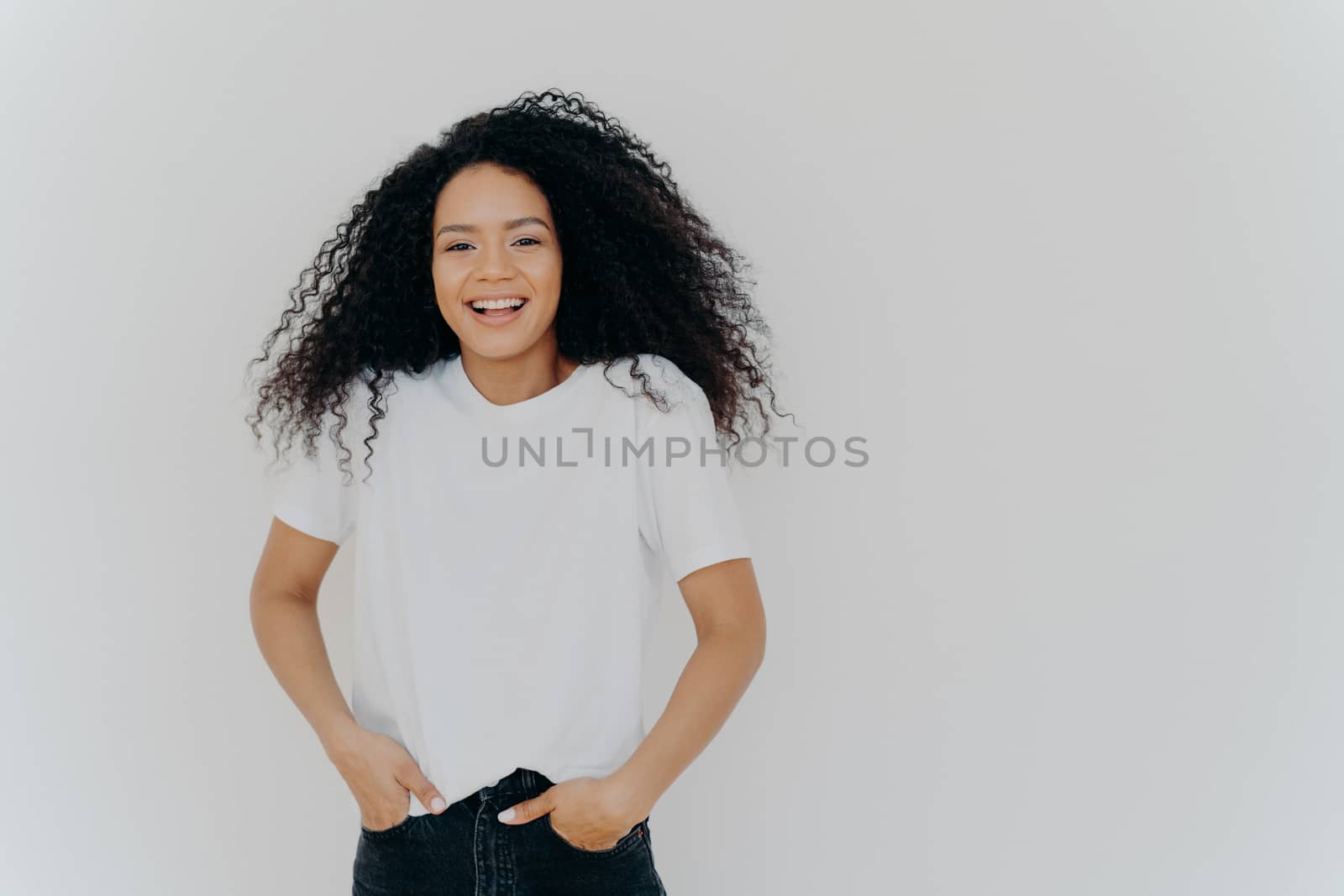 Half length shot of good looking smiling woman laughs at funny joke, has fun, keeps both hands in pockets of jeans, wears white t shirt, has curly fluffy hair, poses indoor, blank space on right side by vkstock