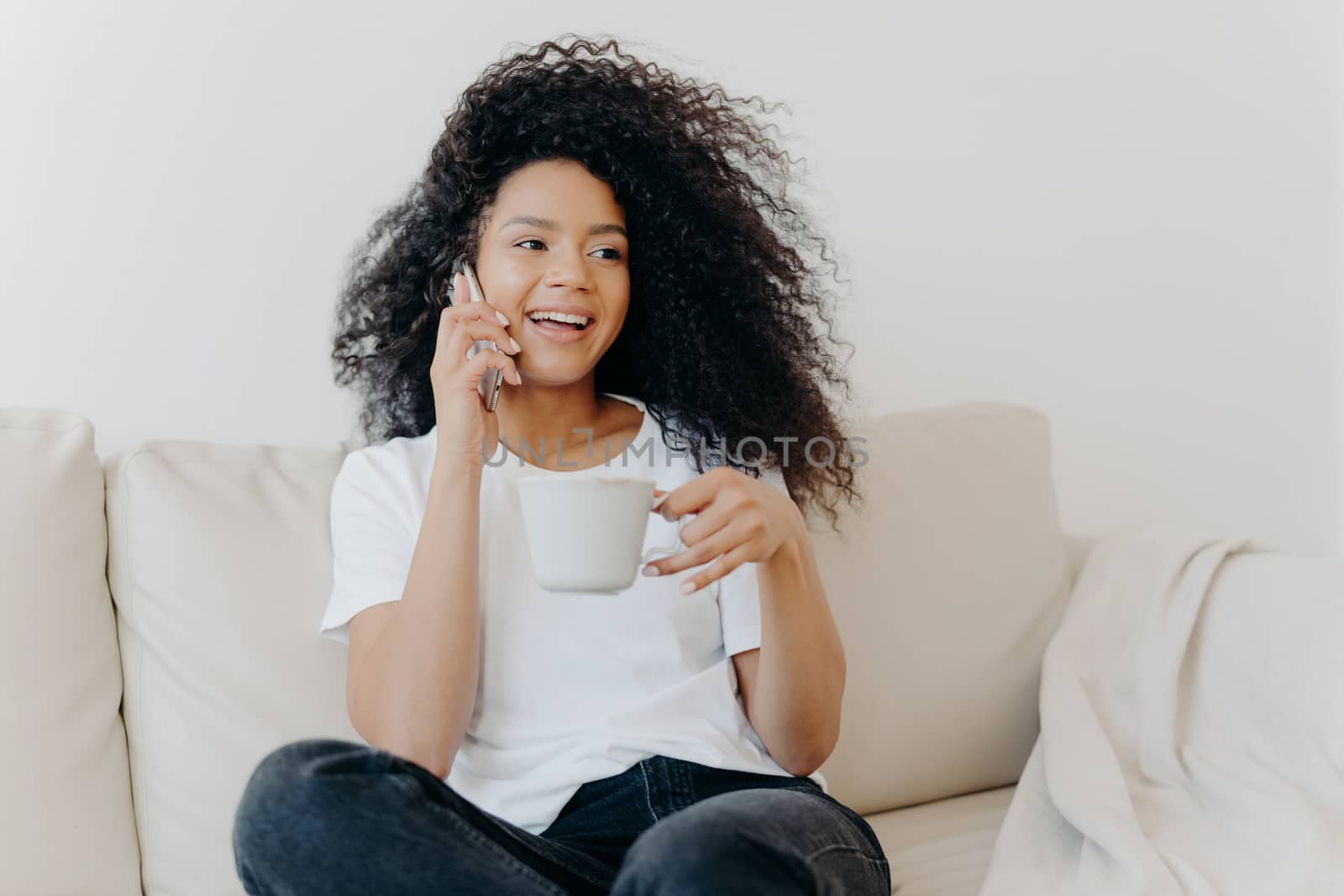 Smiling millennial girl with Afro hair, enjoys cellphone conversation with hot drink in cup, has healthy skin, dressed casually, sits on white sofa at home in modern apartment, orders delivery by vkstock