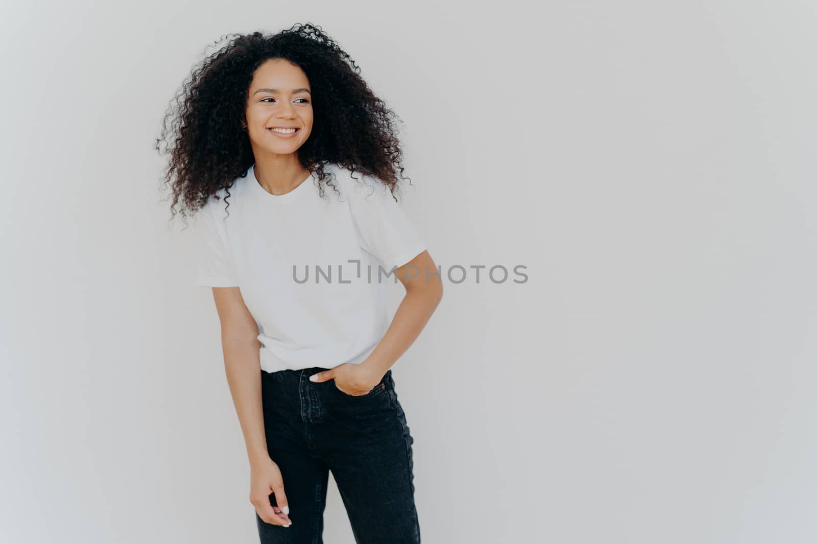 Young curly haired lady has slim figure, wears white t shirt and jeans, looks aside with happy expression, shows healthy teeth, notices funny scene on right side, isolated on white background by vkstock