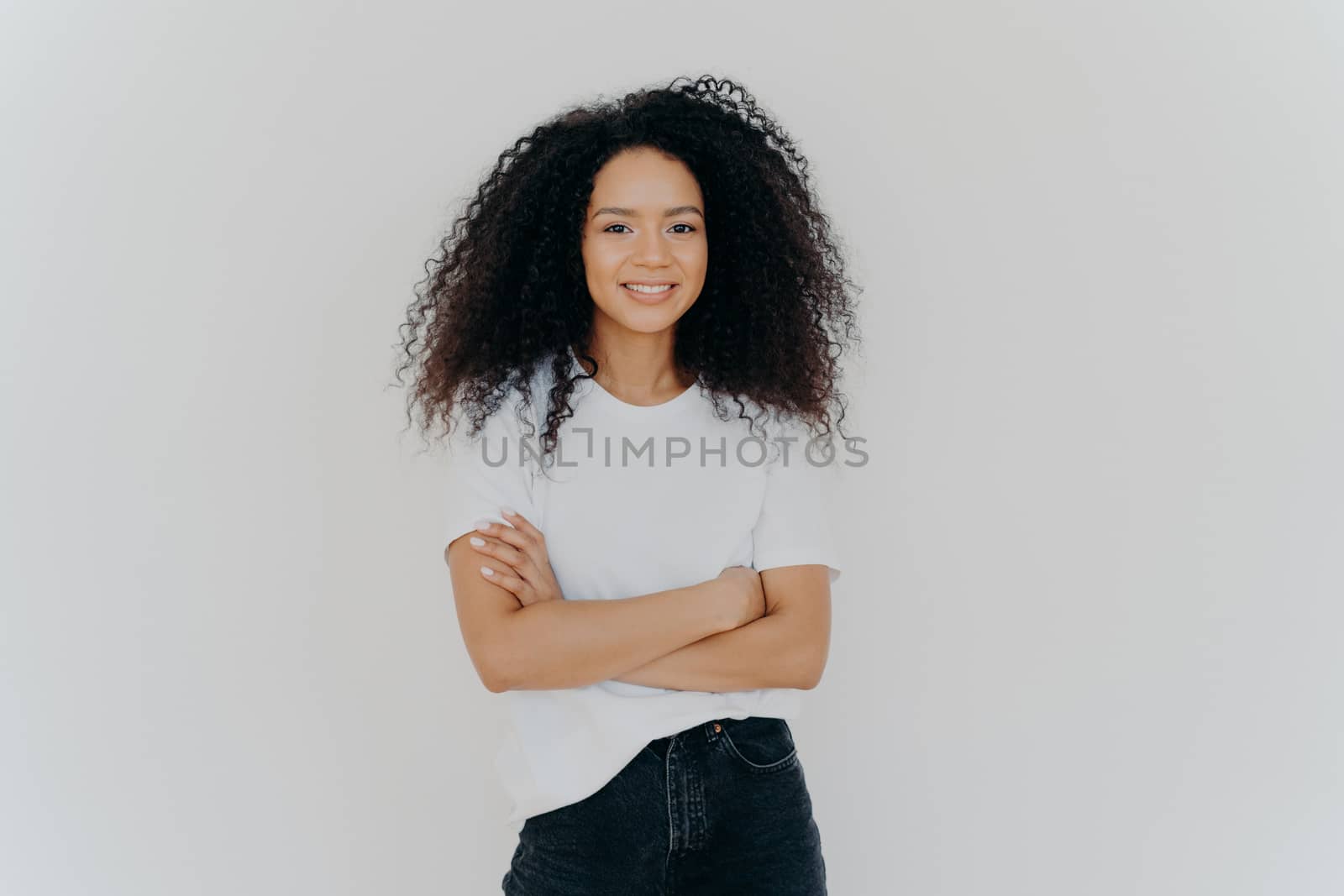 Studio shot of carefree young beautiful woman with Afro hairstyle, keeps arms folded, smiles joyfully, wears casual t shirt and jeans, isolated on white background. People, ethnicity, face expressions by vkstock