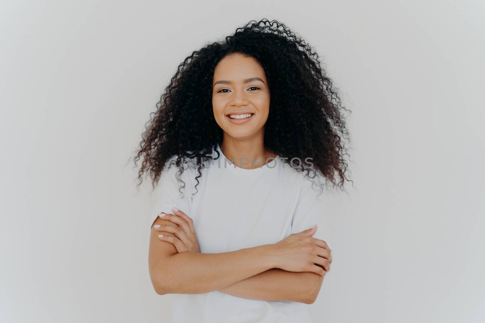 Portrait of young pleasant looking woman with curly hair, keeps hands crossed, wears white t shirt, smiles broadly at camera, has timid shy expression, poses indoor, has natural beauty. Emotions by vkstock