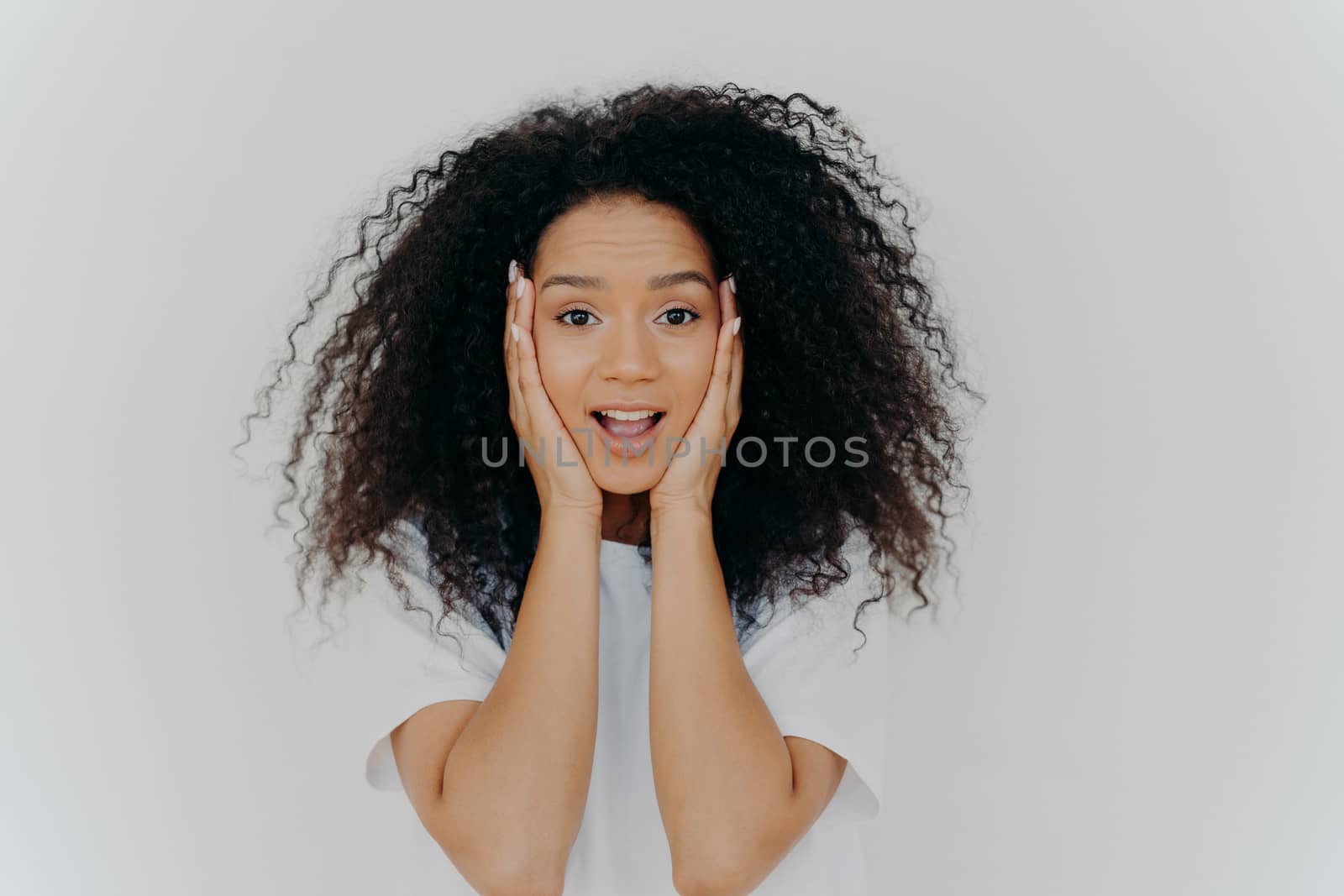Photo of surprised cheerful woman with Afro haircut, keeps both hands on cheeks, has natural beauty, opens mouth, cannot believe in exciting news, wears t shirt, poses against white background. by vkstock