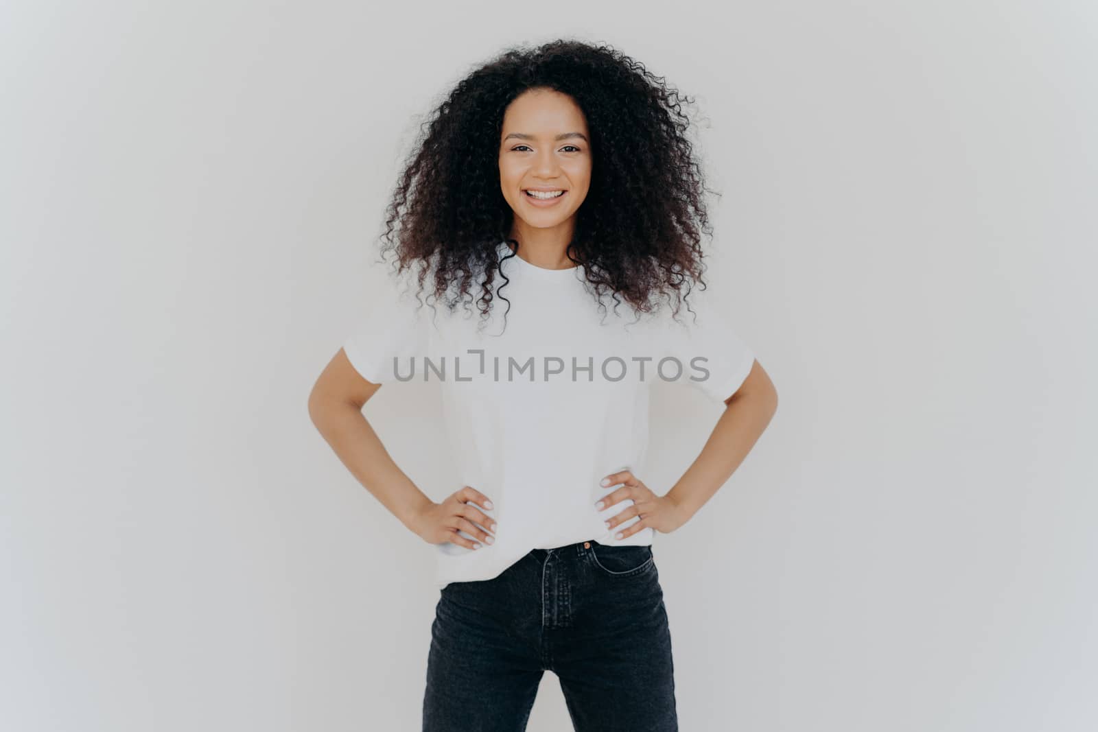 Photo of delighted curly woman keeps both hands on waist, smiles gently, has slim figure, wears white t shirt and black jeans, being in good mood, stands self assured against white background by vkstock