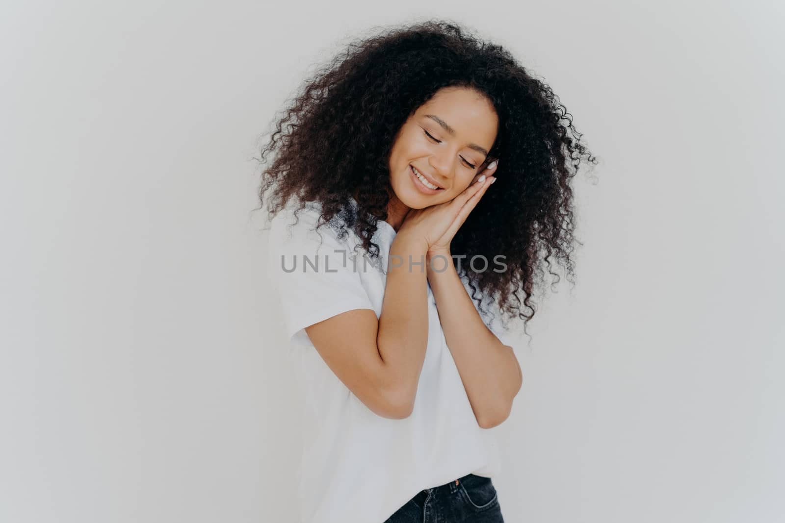 Charming beautiful woman with tender smile, leans on pressed palms, dreams about sleep, tilts head, wears white t shirt, has natural beauty, perfect teeth, poses indoor. Young dreamy Afro woman by vkstock