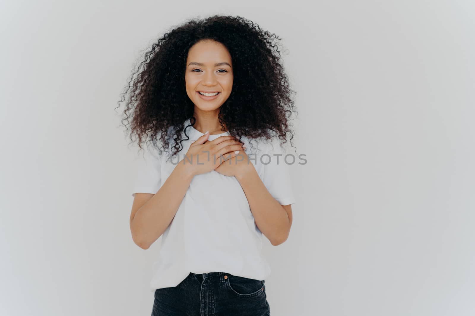 Photo of cheerful woman with Afro hair, keeps hands on chest, expresses gratitude, smiles gently, wears white t shirt and jeans, impressed to get pleasant compliment, isolated over white background by vkstock