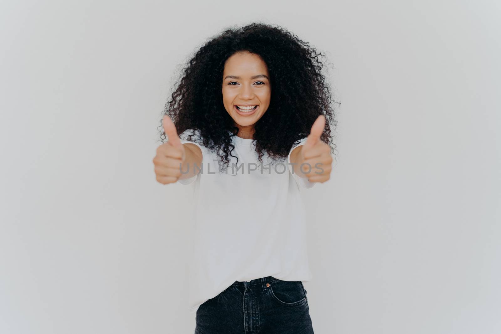 Happy young woman with Afro hairstyle, shows thumbs up, accepts or agrees awesome plan, gives her approval, smiles broadly, stands indoor, says excellent decision, isolated over white background by vkstock