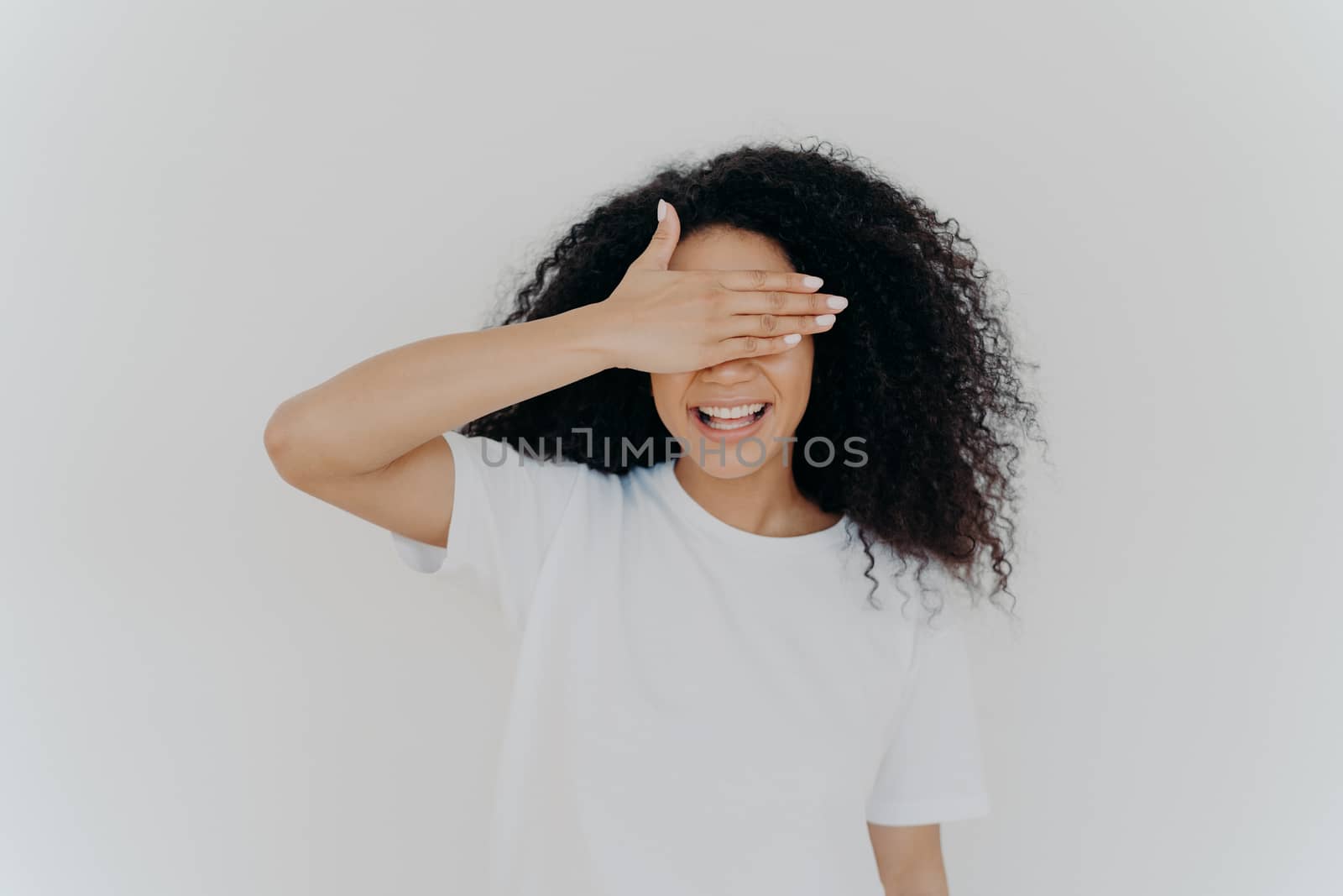 Young teen lady covers eyes with palm, hides herself, smiles toothy, has Afro bushy hairstyle, wears white mockup t shirt, stands alone in studio, waits for surprise, excited from anticipation by vkstock
