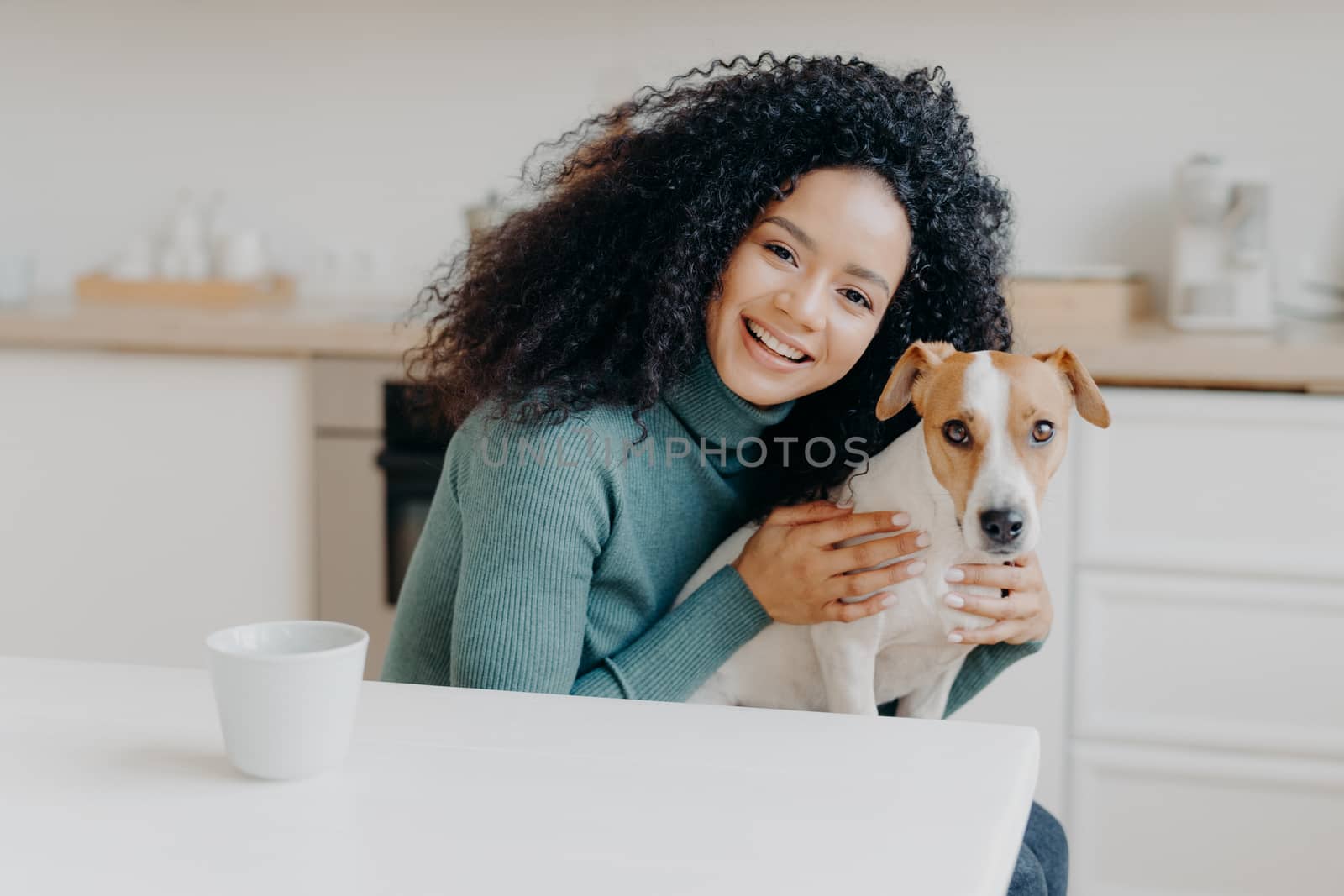 Adorable smiling woman embraces her favourite dog, expresses care and love, good attitiude to pet, poses against kitchen interior, drinks aromatic beverage. People, animals, relationship, affection by vkstock