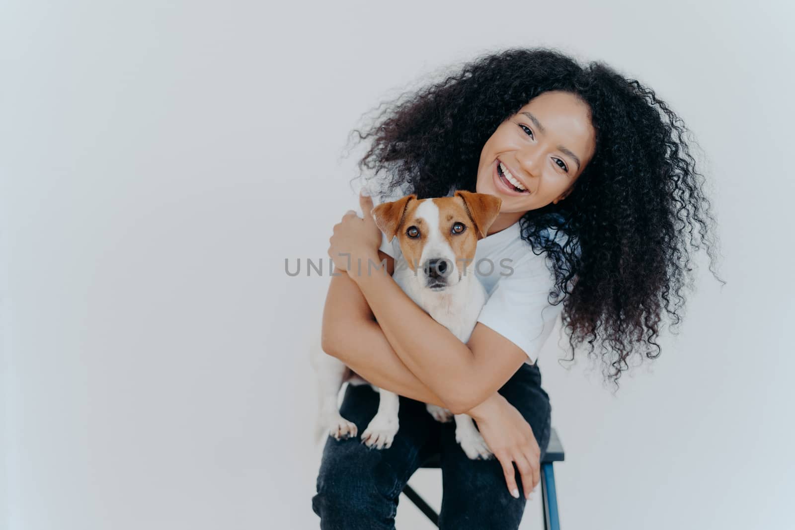 Pleasant looking curly girl tilts head, smiles happily, embraces favourite dog, has good time with pet, wears casual t shirt and jeans, sits on comfortable chair against white background. Relationship