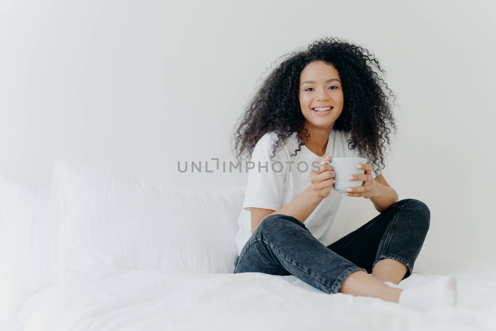 Young pretty African American woman rests in bed at home, drinks hot tea in morning, enjoys domestic atmosphere, tasty drink while relaxes in bedroom. People, leisure, rest and lifestyle concept by vkstock