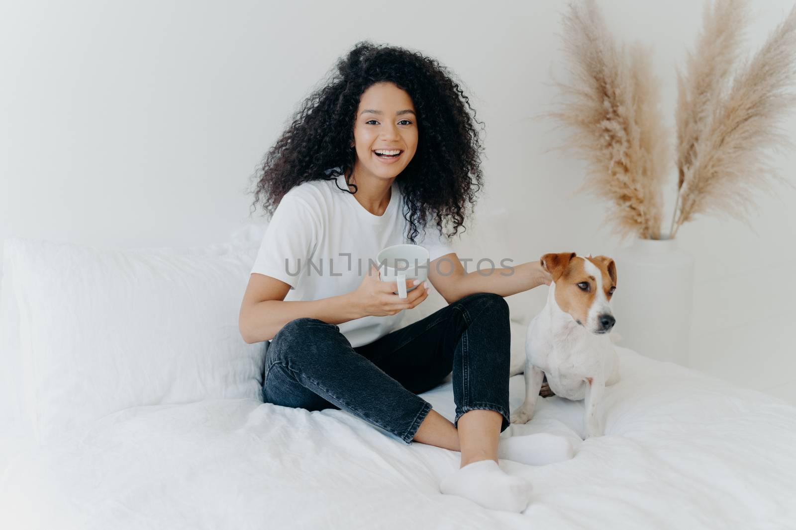 Happy relaxed lovely young woman with Afro hairstyle, petting dog, sit together on bed with white bedclothes, drink hot beverage, dressed in casual wear, expresses positive emotions. People and rest