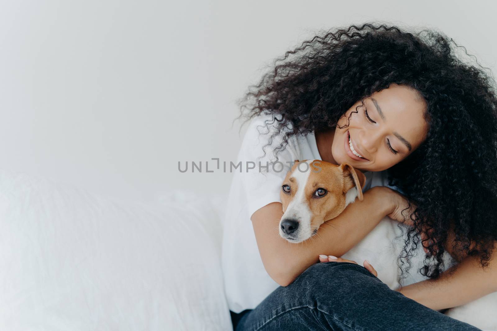 Photo of attractive woman with curly Afro hairstyle, cuddles and pets dog with smile, expresses love, enjoys cozy domestic atmosphere, pose against white background with empty space for promotion