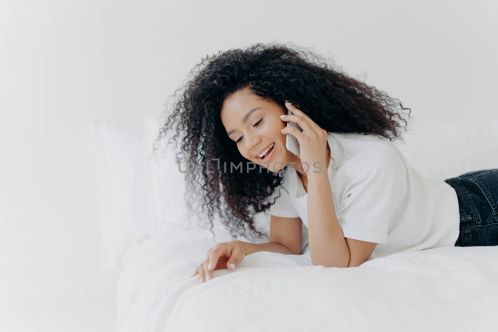 Pleased relaxed woman with curly hair has pleasant telephone conversation, speaks with friend during weekend before sleep, wears white t shirt, lies in cozy bed, keeps modern gadget near ear by vkstock