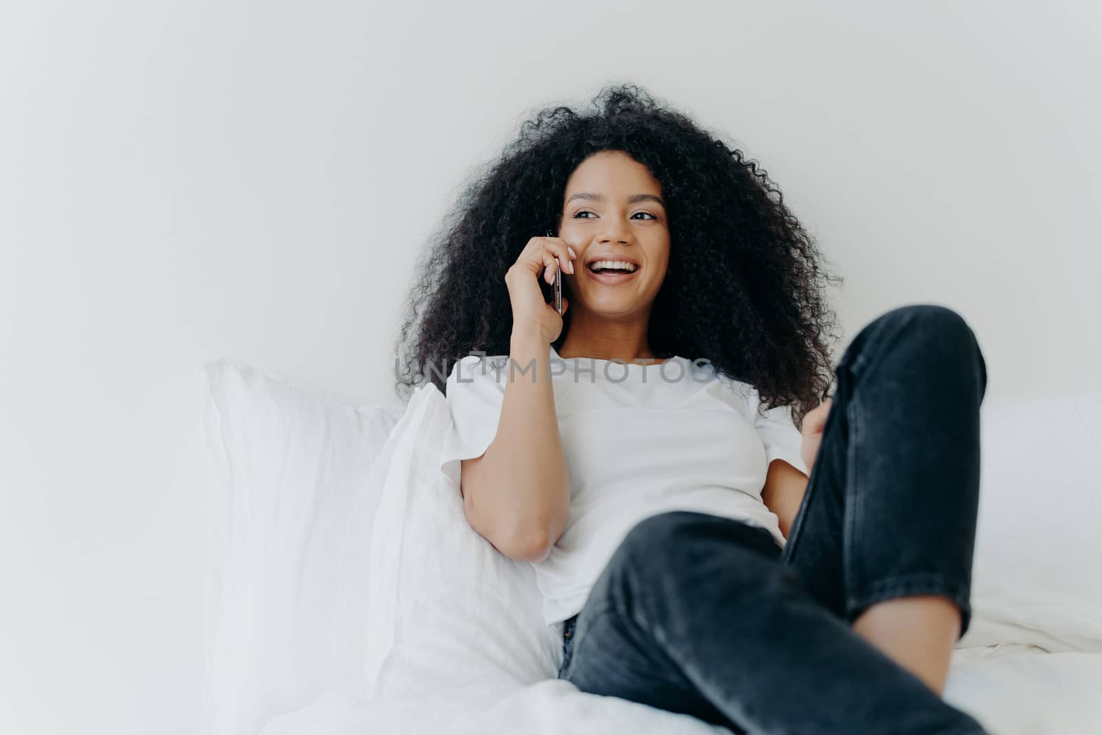 Pleasant looking curly woman has pleasant conversation, talks with friend via smartphone while relaxes in bed, has joyful expression, looks aside, wears casual clothing. People, bedding, technology