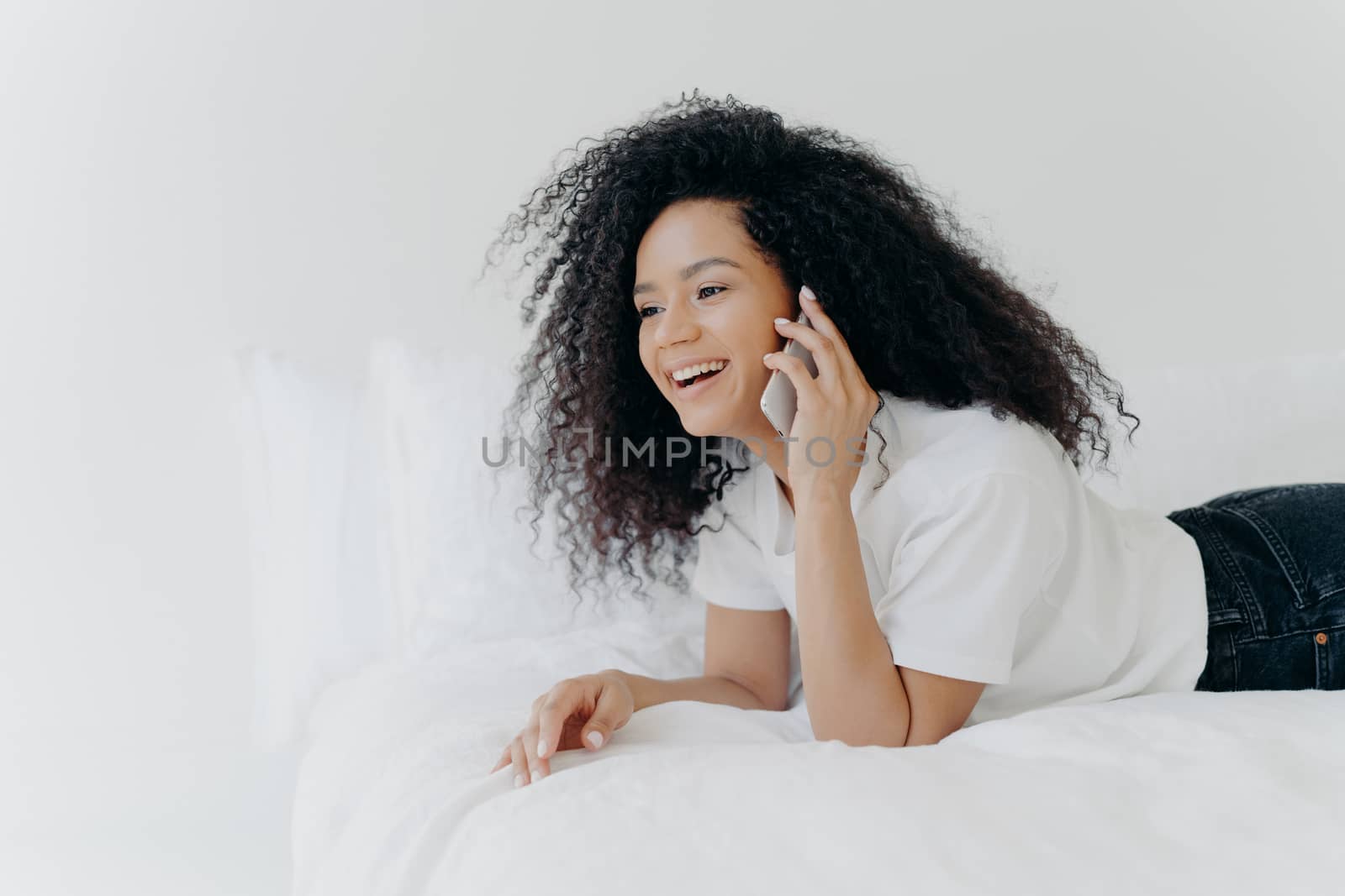 Charming young woman with crisp hair, speaks with boyfriend via smartphone, has rest in morning at bed, laughs at funny joke, dressed casually, being at home against white wall. Communication concept