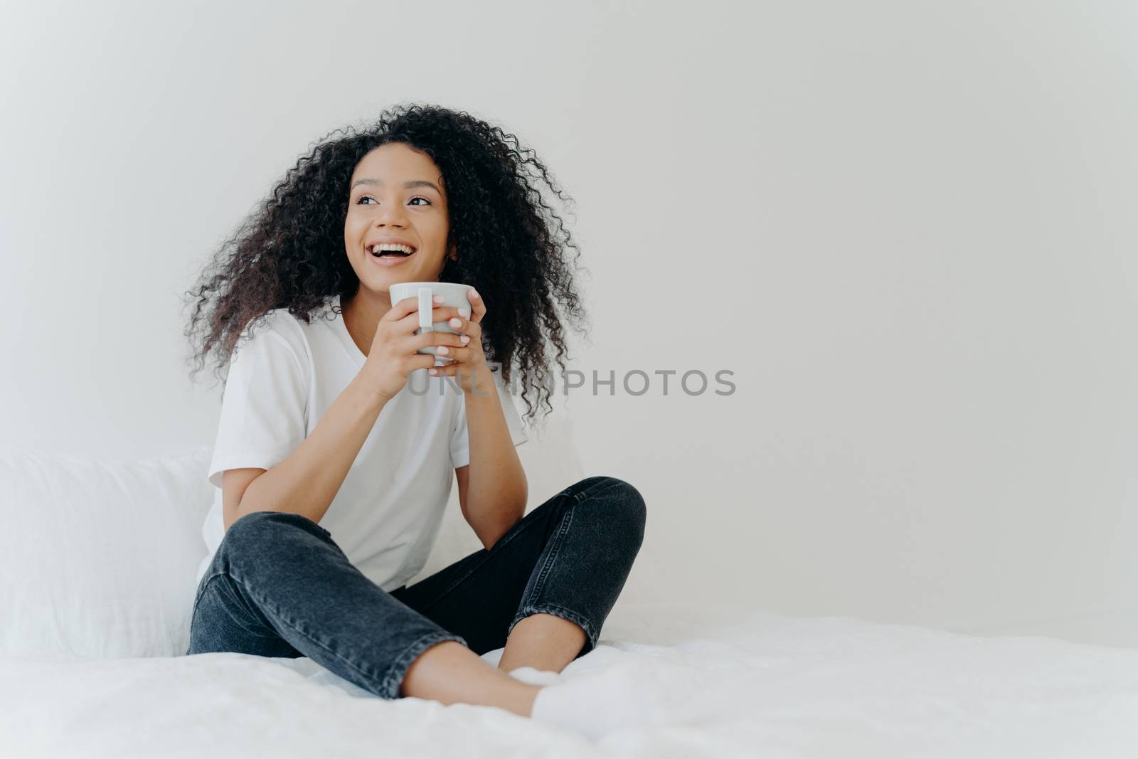Lazy morning and bedtime concept. Positive Afro woman holds mug of hot tea or coffee, sits in bed, looks gladfully aside, enjoys good weather, wears comfortable clothes, white wall in background by vkstock