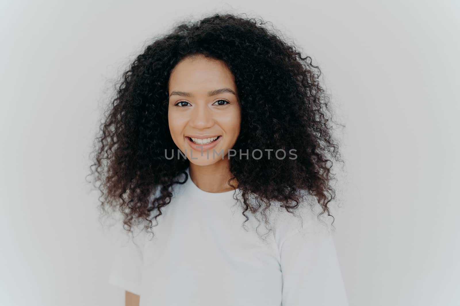 Happiness, wellbeing and positive emotions concept. Headshot of pleasant looking lady with crisp hair, smiles and laughs, has healthy skin, wears casual t shirt, models against white studio wall. by vkstock
