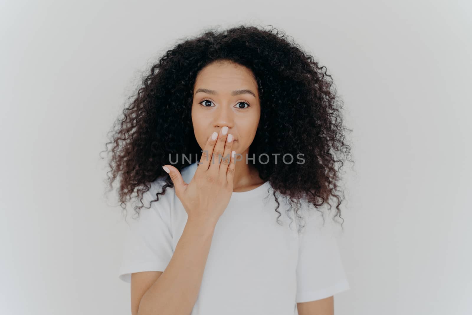 Headshot of impressed curly young woman covers mouth with palm, tries to be speechless, has curly hairstyle, manicure and minimal makeup, wears white t shirt, poses indoor. Omg, what gossip! by vkstock