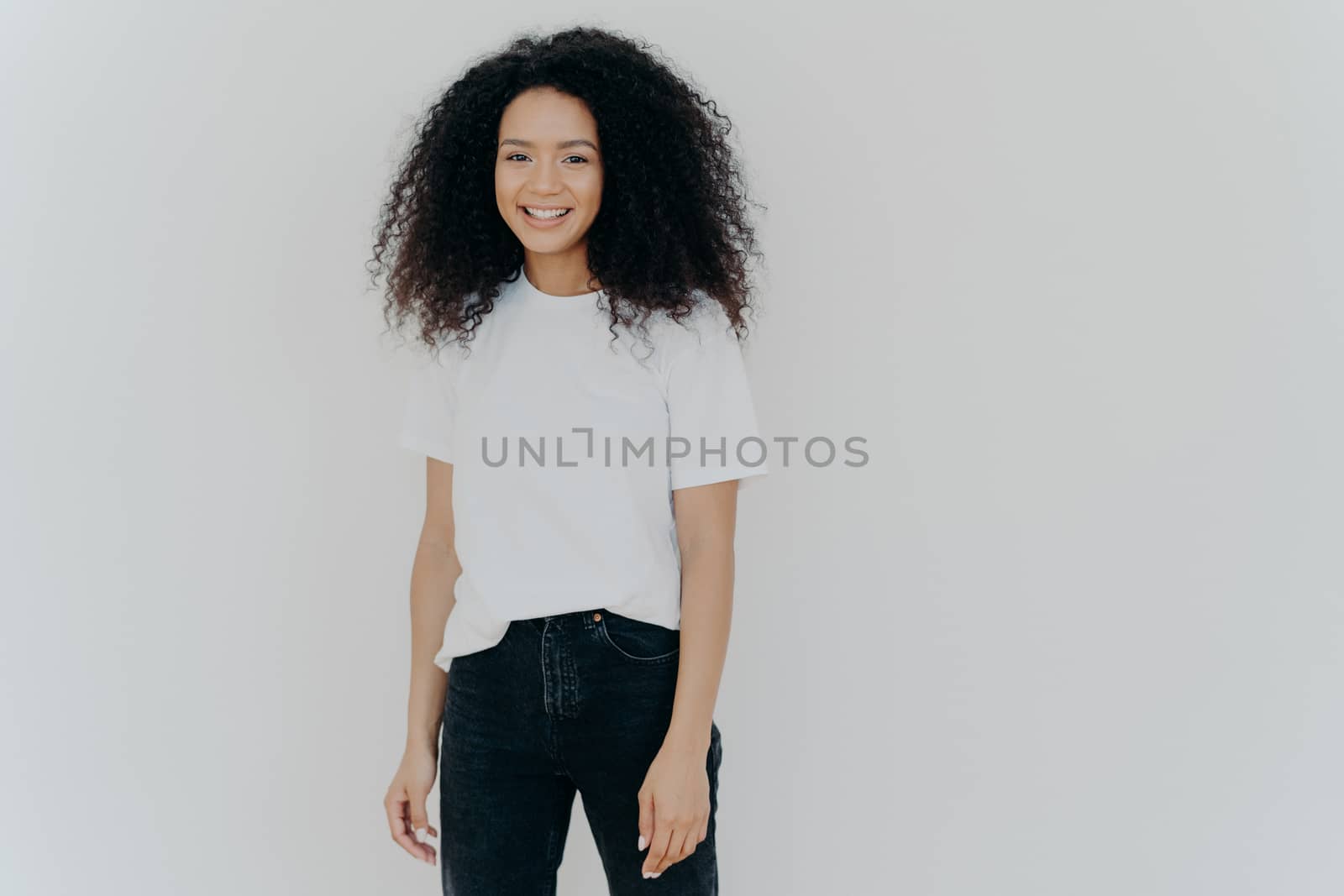 Picture of good looking woman with gentle smile, stands satisfied, has sincere carefree expression, relaxed casual mood, dressed in casual tshirt and black jeans, isolated over white background by vkstock