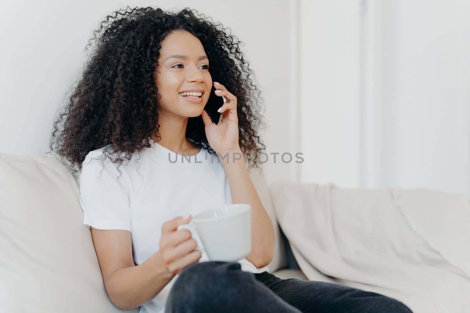 Sideways shot of positive ethnic brunette woman has telephone conversation, drinks hot beverage, holds white cup, sits on comfortable couch in living room, hears pleasant news, smiles happily by vkstock