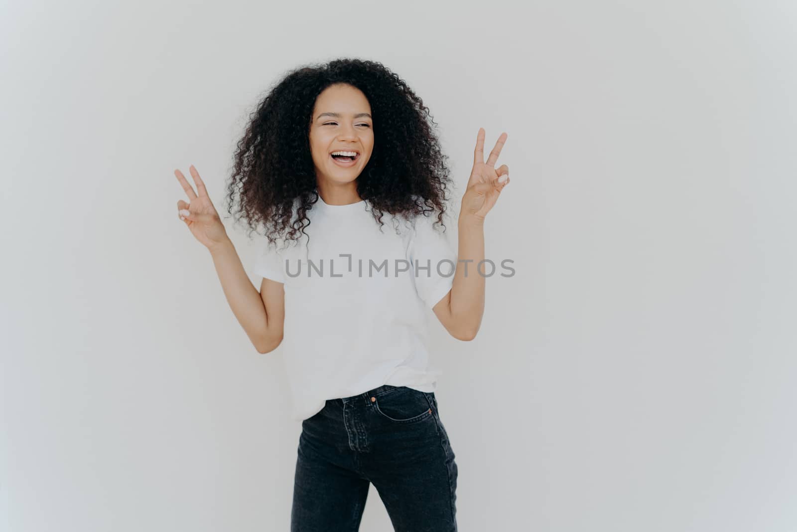 Carefree woman with Afro hairstyle raises hands up, shows victory gesture or peace sign, looks gladfully aside, dressed in casual wear, smiles relaxed, sends hello, isolated over white background.